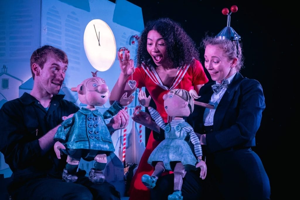 Three performers on stage interact with two large puppets. A man in black operates a puppet of a boy in a blue coat, while a woman in a suit and bow tie with a fun hat shaped like an antenna operates a puppet of a girl in a blue dress. Between them, a woman in a bright red dress holds up a shiny silver heart, smiling as the puppets reach toward it. A clock showing nearly midnight and candy canes decorate the background.