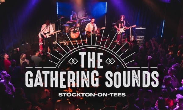 Live music venue photograph showing a band performing on stage with dramatic purple and blue stage lighting. The stage features a four-piece band with guitars, drums, and other instruments, with atmospheric smoke effects. In the foreground, a large crowd of audience members can be seen enjoying the performance, many with raised hands. Overlaid on the image is 'The Gathering Sounds' logo in large white distressed text with a decorative sunburst design above it, and 'Stockton-on-Tees' underneath. The scene captures the energetic atmosphere of a live music event in an intimate venue setting