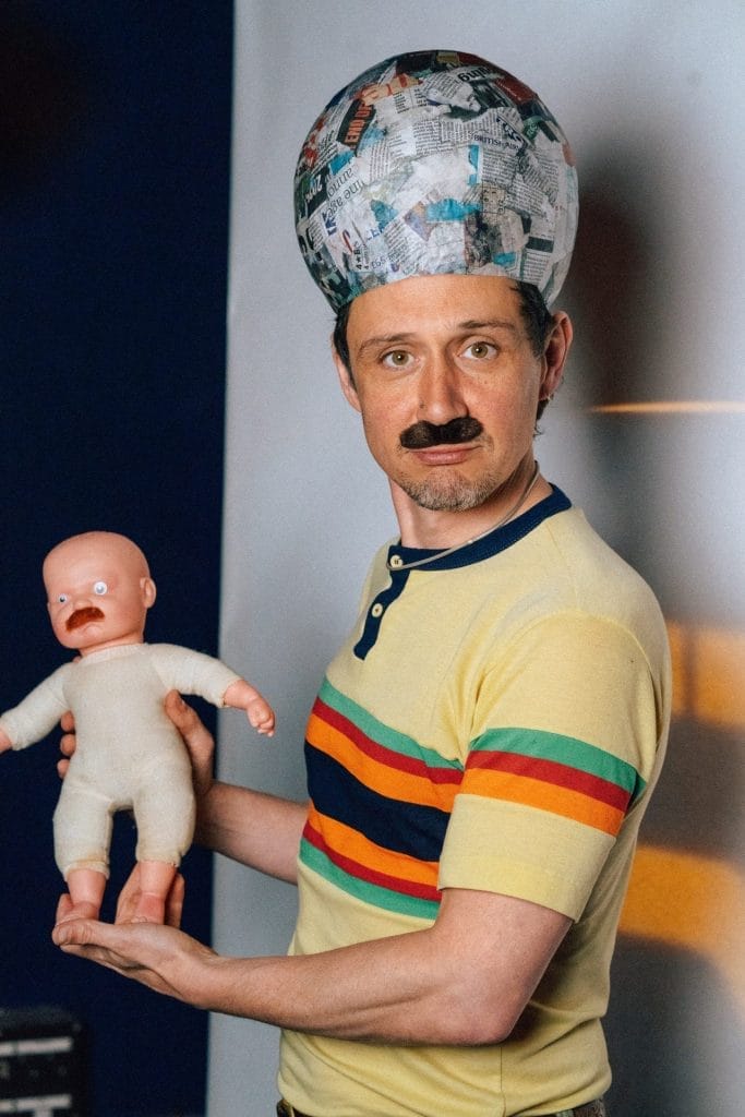 A man wearing a yellow retro-style shirt with colorful stripes holds a baby doll. He has a fake black mustache and is wearing a large papier-mâché hat made of newspaper clippings. The doll also has a mustache. The background is split into dark blue and white sections.