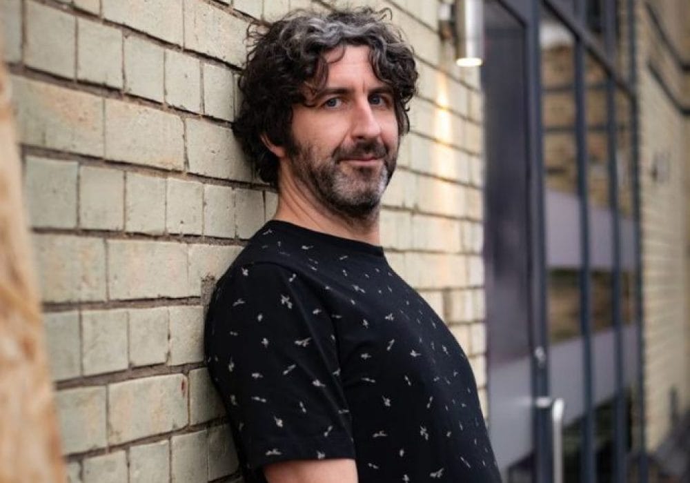 An Image of mark Watson leaning against a brick wall. He has mid-length dark hair and a beard, he is wearing a dark blue patterned top and is looking directly at the camera.