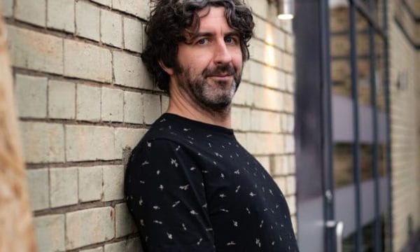 An Image of mark Watson leaning against a brick wall. He has mid-length dark hair and a beard, he is wearing a dark blue patterned top and is looking directly at the camera.