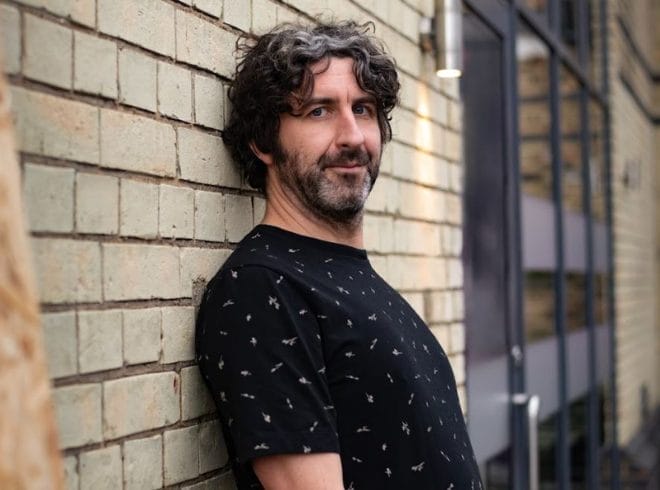 An Image of mark Watson leaning against a brick wall. He has mid-length dark hair and a beard, he is wearing a dark blue patterned top and is looking directly at the camera.