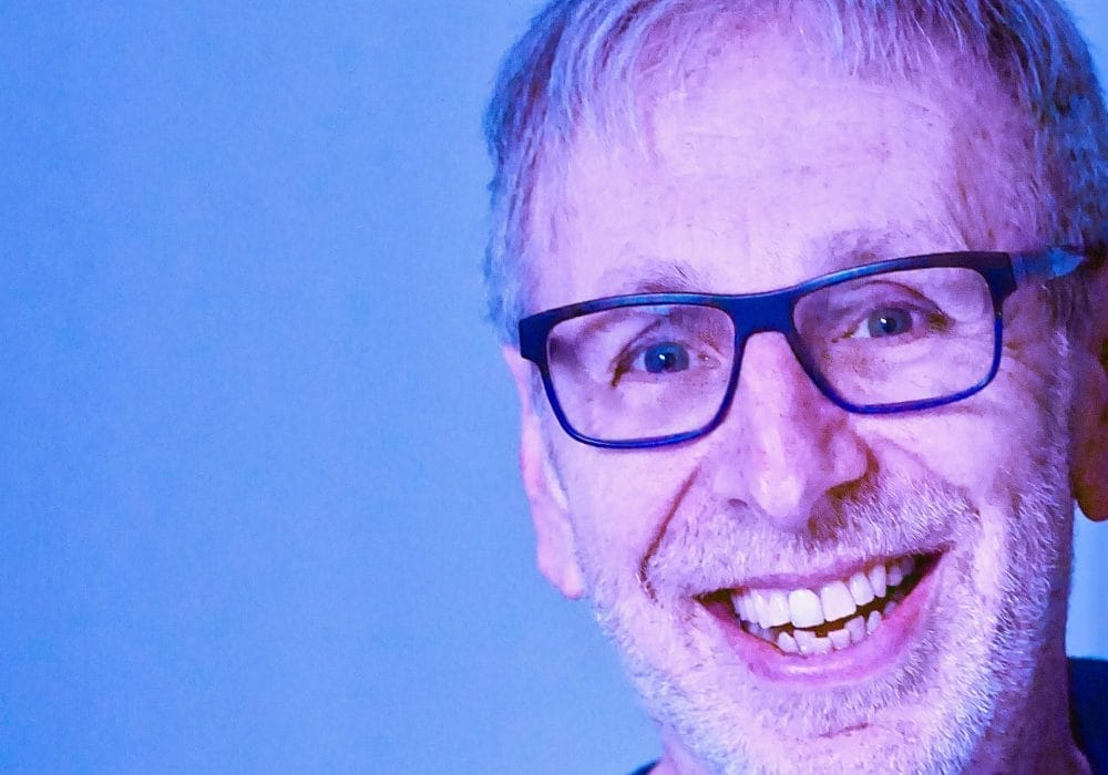 An image of a man smiling wearing glasses and a black shirt. there is a blue background