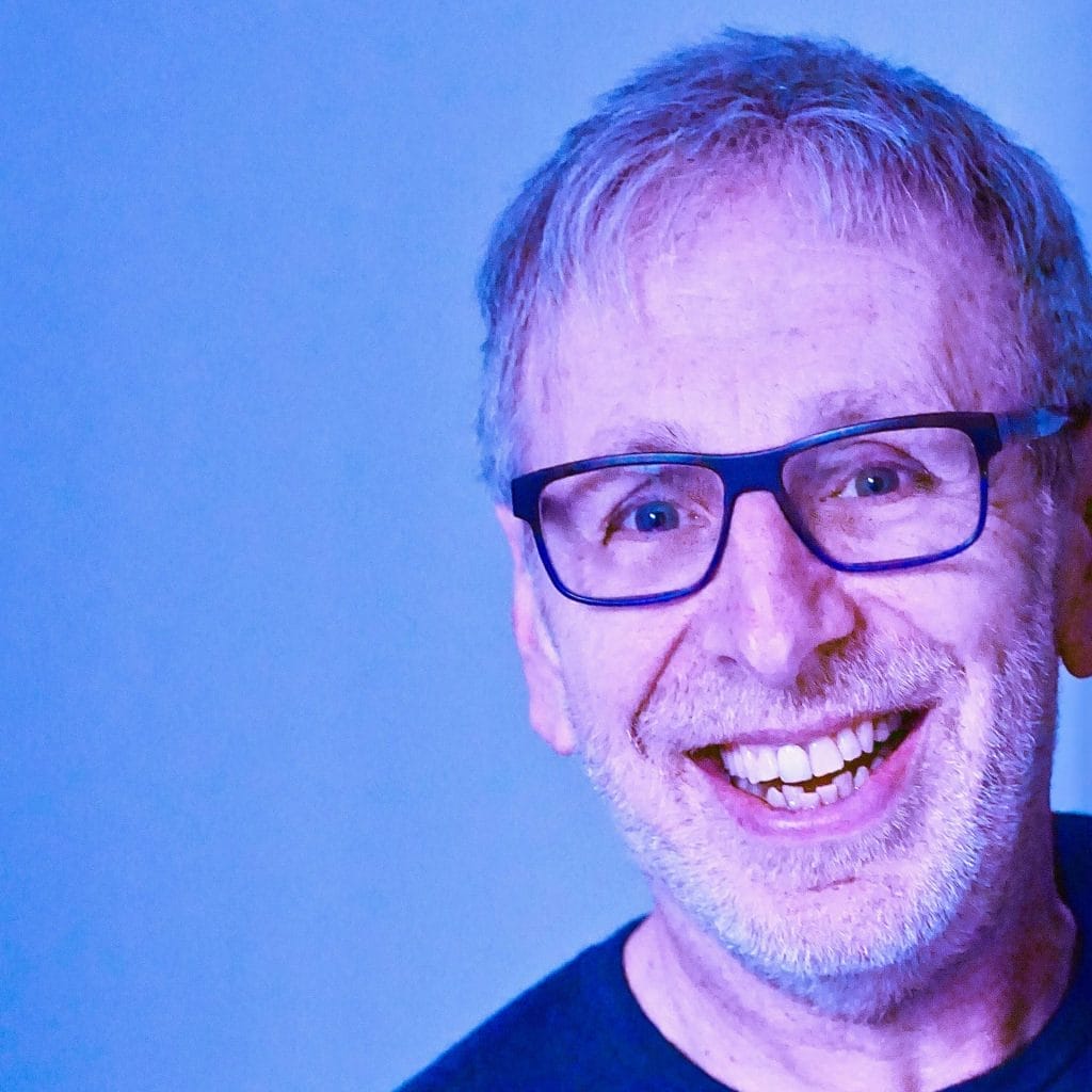An image of a man smiling wearing glasses and a black shirt. there is a blue background