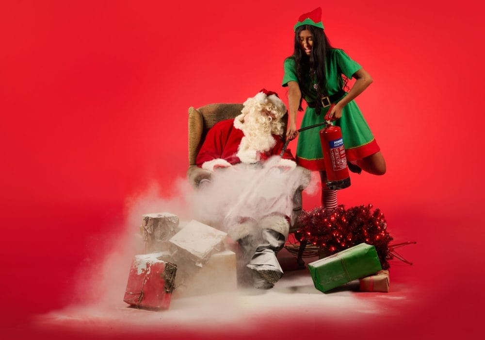 Santa sat sleeping in a chair while an elf is using a fire extinguisher to put out a fire beside him.