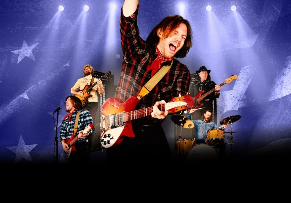 A dynamic concert scene featuring a rock band performing on stage with dramatic blue and purple stage lighting. The lead guitarist in the foreground is energetically playing a red and white electric guitar while wearing a black and red plaid shirt, with one arm raised triumphantly. Behind him, other band members are visible including a bassist, drummer, and additional guitarists, all performing against a backdrop decorated with star patterns. The stage is illuminated by multiple spotlights creating a vibrant concert atmosphere