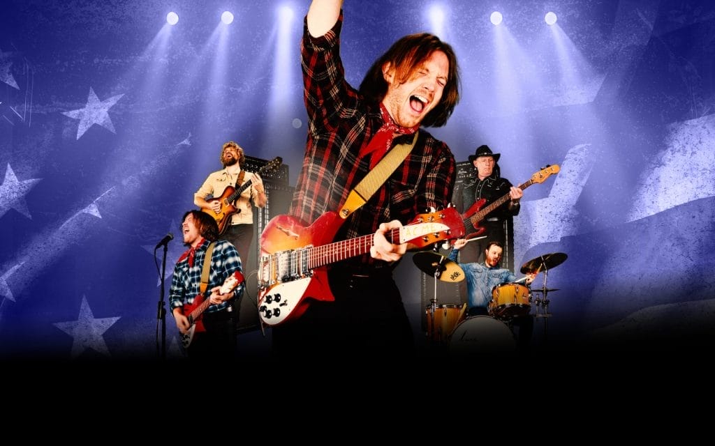 A dynamic concert scene featuring a rock band performing on stage with dramatic blue and purple stage lighting. The lead guitarist in the foreground is energetically playing a red and white electric guitar while wearing a black and red plaid shirt, with one arm raised triumphantly. Behind him, other band members are visible including a bassist, drummer, and additional guitarists, all performing against a backdrop decorated with star patterns. The stage is illuminated by multiple spotlights creating a vibrant concert atmosphere