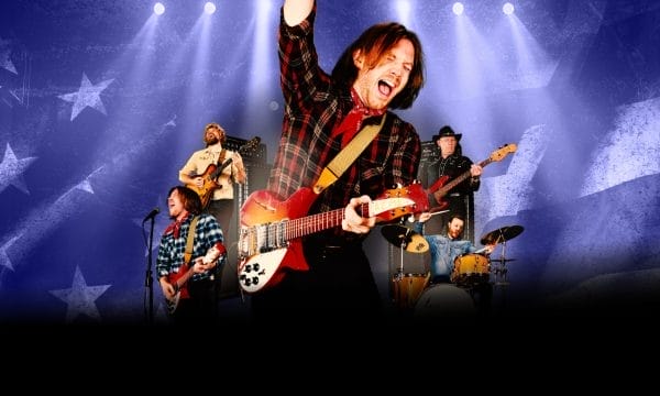 A dynamic concert scene featuring a rock band performing on stage with dramatic blue and purple stage lighting. The lead guitarist in the foreground is energetically playing a red and white electric guitar while wearing a black and red plaid shirt, with one arm raised triumphantly. Behind him, other band members are visible including a bassist, drummer, and additional guitarists, all performing against a backdrop decorated with star patterns. The stage is illuminated by multiple spotlights creating a vibrant concert atmosphere