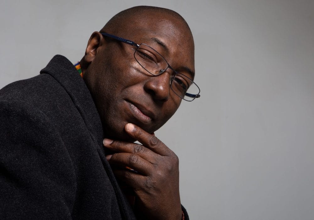 A Black man wearing glasses and what appears to be a dark jumper or hoodie. He's positioned in a thoughtful pose with his hand near his chin