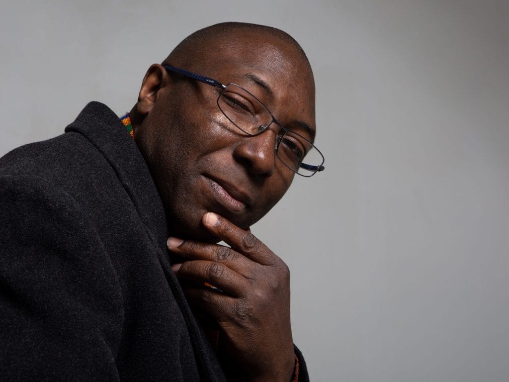 A Black man wearing glasses and what appears to be a dark jumper or hoodie. He's positioned in a thoughtful pose with his hand near his chin