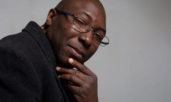 A Black man wearing glasses and what appears to be a dark jumper or hoodie. He's positioned in a thoughtful pose with his hand near his chin
