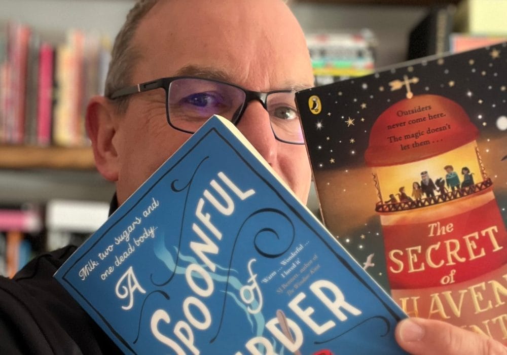 A man with glasses holding up a blue book titled "A Spoonful of Murder" in front of his face, with bookshelves visible in the background. There's also what appears to be another book called "The Secret of [something]" visible in the background.