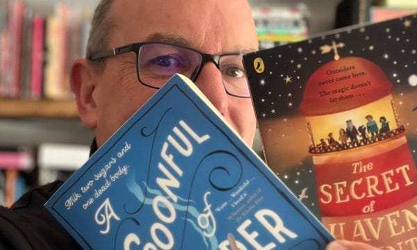 A man with glasses holding up a blue book titled "A Spoonful of Murder" in front of his face, with bookshelves visible in the background. There's also what appears to be another book called "The Secret of [something]" visible in the background.