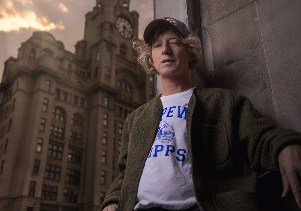 A white man in a cap stands against a wall with a clock tower behind him