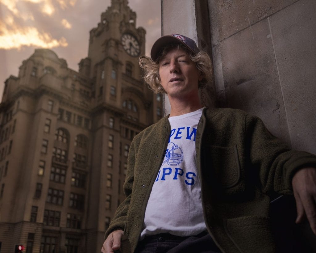A white man in a cap stands against a wall with a clock tower behind him