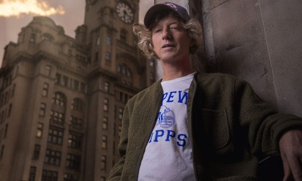 A white man in a cap stands against a wall with a clock tower behind him