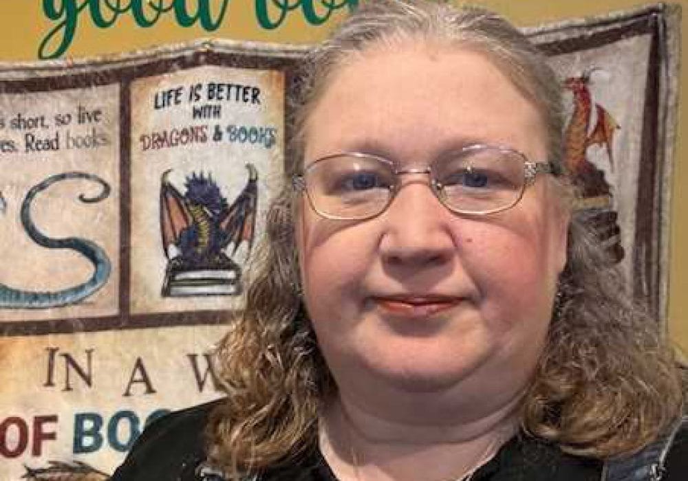 A woman with glasses and shoulder-length hair, sitting in what looks like a room with book-themed decorations or posters on the wall behind her. The setting suggests a bookish or literary environment.