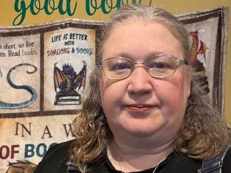 A woman with glasses and shoulder-length hair, sitting in what looks like a room with book-themed decorations or posters on the wall behind her. The setting suggests a bookish or literary environment.