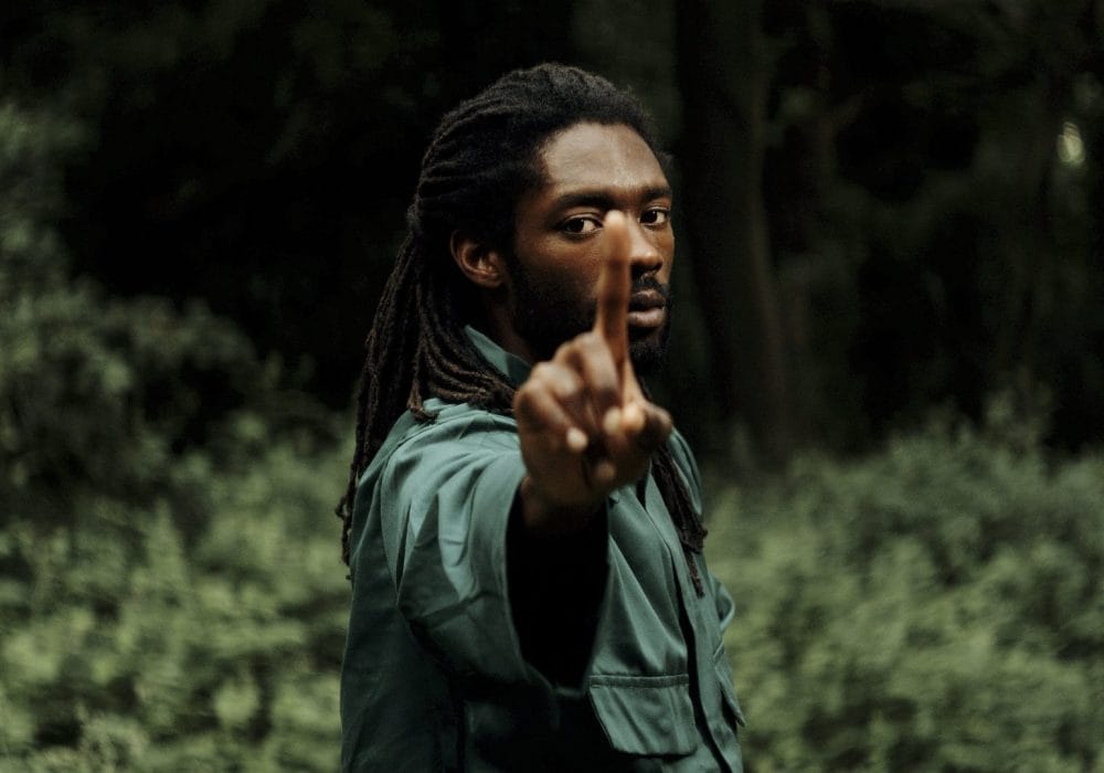 A man stood holding his index finger up towards the screen, he is wearing a green top with long dark hair, the background is outside with trees.