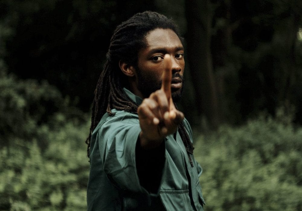 A man stood holding his index finger up towards the screen, he is wearing a green top with long dark hair, the background is outside with trees.