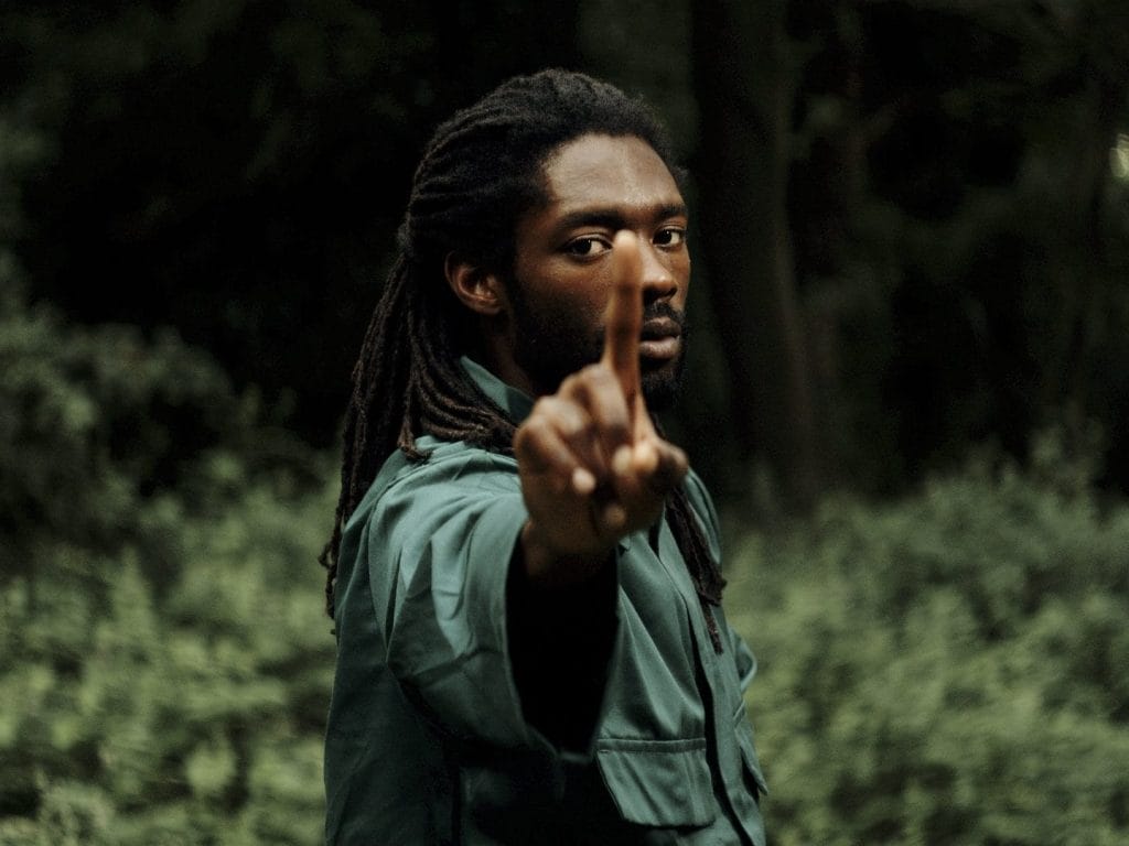 A man stood holding his index finger up towards the screen, he is wearing a green top with long dark hair, the background is outside with trees.