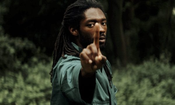A man stood holding his index finger up towards the screen, he is wearing a green top with long dark hair, the background is outside with trees.