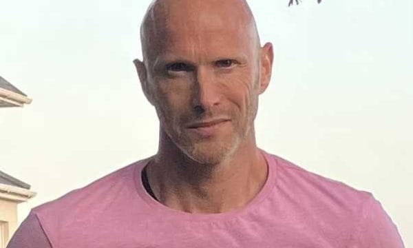 A man in a pink/salmon coloured t-shirt standing outdoors, with what appears to be a building structure in the background. He's bald with facial hair and appears to be in a sunny location.