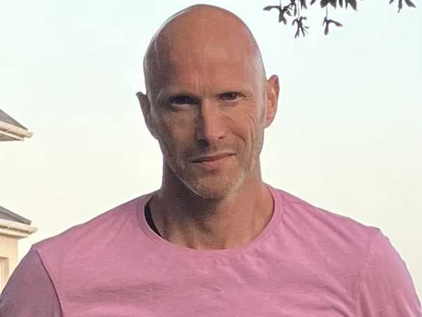 A man in a pink/salmon coloured t-shirt standing outdoors, with what appears to be a building structure in the background. He's bald with facial hair and appears to be in a sunny location.