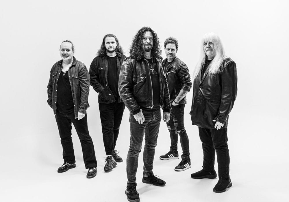a black and white photo of five musicians in a classic rock/metal band formation. They're wearing dark clothing - leather jackets, dark jeans, and casual attire that's typical of rock musicians.