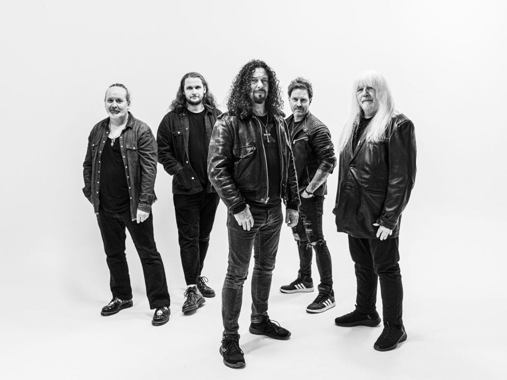 a black and white photo of five musicians in a classic rock/metal band formation. They're wearing dark clothing - leather jackets, dark jeans, and casual attire that's typical of rock musicians.