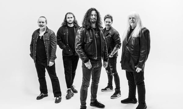 a black and white photo of five musicians in a classic rock/metal band formation. They're wearing dark clothing - leather jackets, dark jeans, and casual attire that's typical of rock musicians.
