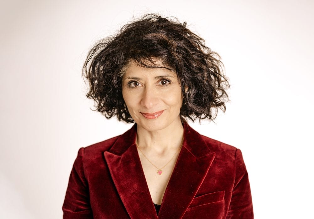 Person with curly dark hair wearing a red velvet blazer and a necklace, smiling softly against a plain light background.