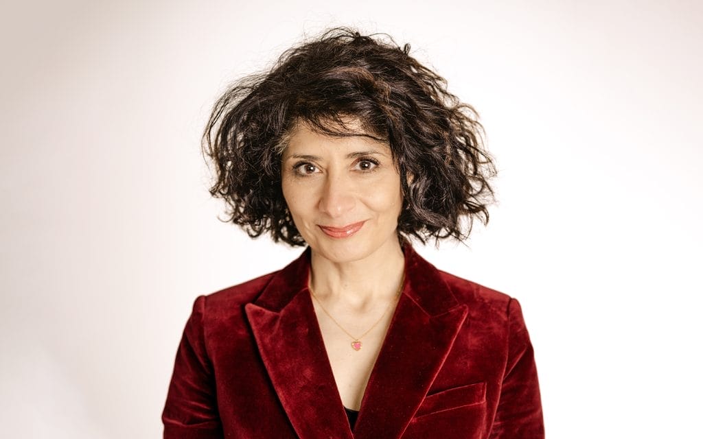 Person with curly dark hair wearing a red velvet blazer and a necklace, smiling softly against a plain light background.