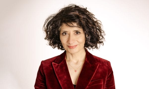 Person with curly dark hair wearing a red velvet blazer and a necklace, smiling softly against a plain light background.
