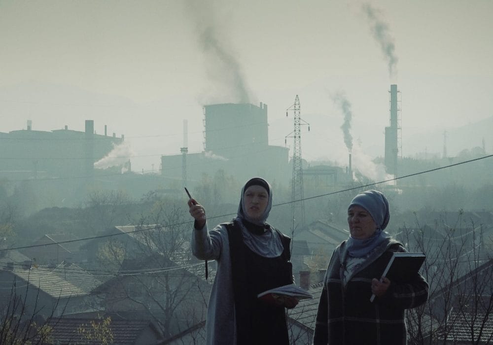 Two women in headscarves stand in front of a polluted industrial landscape, holding notebooks. One gestures with a pen as smoke rises from factories behind them.