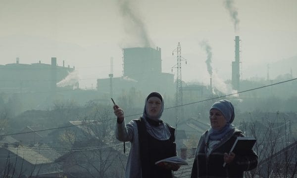 Two women in headscarves stand in front of a polluted industrial landscape, holding notebooks. One gestures with a pen as smoke rises from factories behind them.
