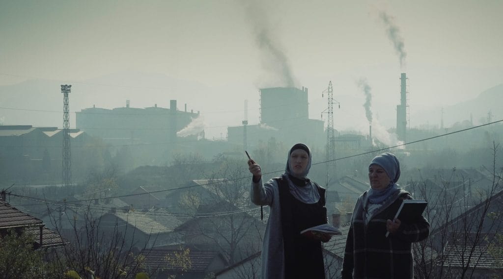 Two women in headscarves stand in front of a polluted industrial landscape, holding notebooks. One gestures with a pen as smoke rises from factories behind them.
