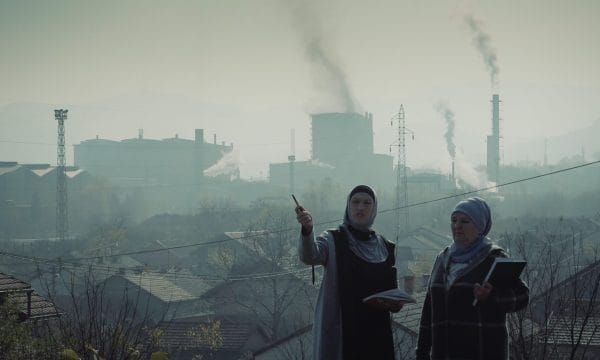 Two women in headscarves stand in front of a polluted industrial landscape, holding notebooks. One gestures with a pen as smoke rises from factories behind them.