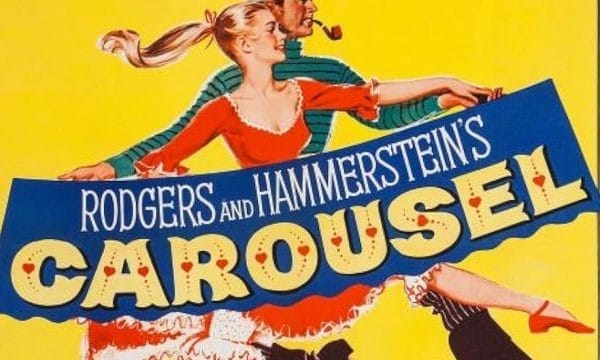 a bright yellow background with a man and woman in 1940s period costume. The woman wears a red dress with white trim and has blonde hair flowing in the wind. The man behind her wears a green and white striped shirt and a flat cap, smoking a pipe. They appear to be running or moving dynamically across the poster. The title "CAROUSEL" is displayed prominently in large, decorative letters with red heart symbols dotting the text. Above the title, "RODGERS AND HAMMERSTEIN'S" appears in white text on a blue banner.