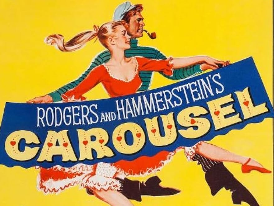 a bright yellow background with a man and woman in 1940s period costume. The woman wears a red dress with white trim and has blonde hair flowing in the wind. The man behind her wears a green and white striped shirt and a flat cap, smoking a pipe. They appear to be running or moving dynamically across the poster. The title "CAROUSEL" is displayed prominently in large, decorative letters with red heart symbols dotting the text. Above the title, "RODGERS AND HAMMERSTEIN'S" appears in white text on a blue banner.