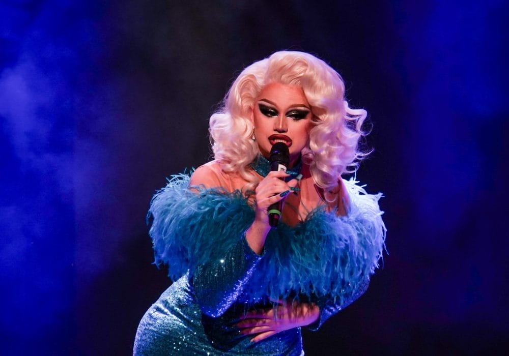 A glamorous drag performer with voluminous platinum blonde curls and dramatic makeup sings into a microphone on stage. They are wearing a sparkling teal dress with feathered detailing on the shoulders, lit by vibrant blue and purple stage lighting