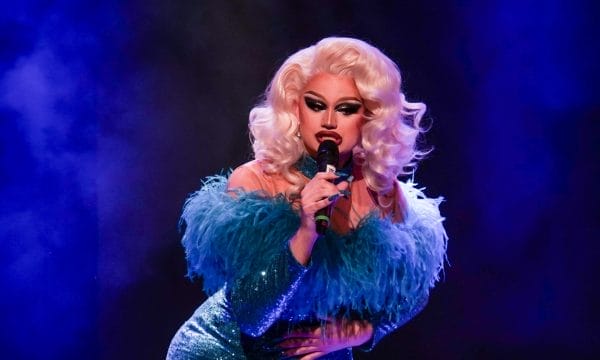 A glamorous drag performer with voluminous platinum blonde curls and dramatic makeup sings into a microphone on stage. They are wearing a sparkling teal dress with feathered detailing on the shoulders, lit by vibrant blue and purple stage lighting