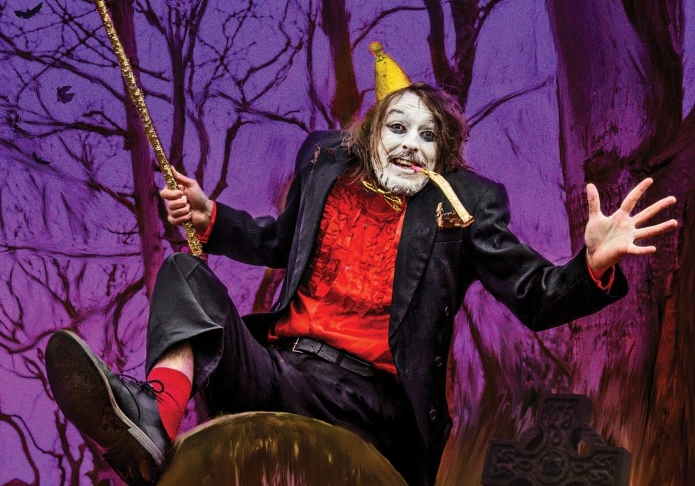 A theatrical performer in elaborate costume sits dramatically on a large mushroom prop against a purple-tinted forest backdrop. They wear a black coat over a red vest, with white face paint, a small yellow party hat, and hold a golden wand or staff. The scene has a whimsical, fairy-tale theatre production aesthetic with bare tree branches visible in the purple-hued background.