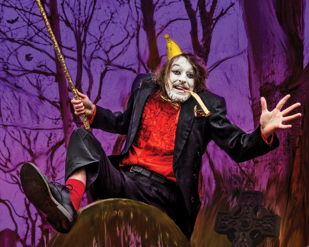 A theatrical performer in elaborate costume sits dramatically on a large mushroom prop against a purple-tinted forest backdrop. They wear a black coat over a red vest, with white face paint, a small yellow party hat, and hold a golden wand or staff. The scene has a whimsical, fairy-tale theatre production aesthetic with bare tree branches visible in the purple-hued background.