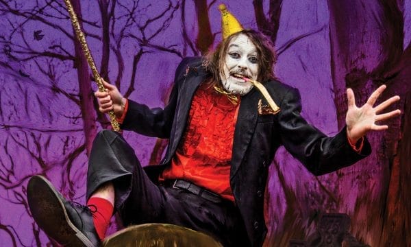 A theatrical performer in elaborate costume sits dramatically on a large mushroom prop against a purple-tinted forest backdrop. They wear a black coat over a red vest, with white face paint, a small yellow party hat, and hold a golden wand or staff. The scene has a whimsical, fairy-tale theatre production aesthetic with bare tree branches visible in the purple-hued background.