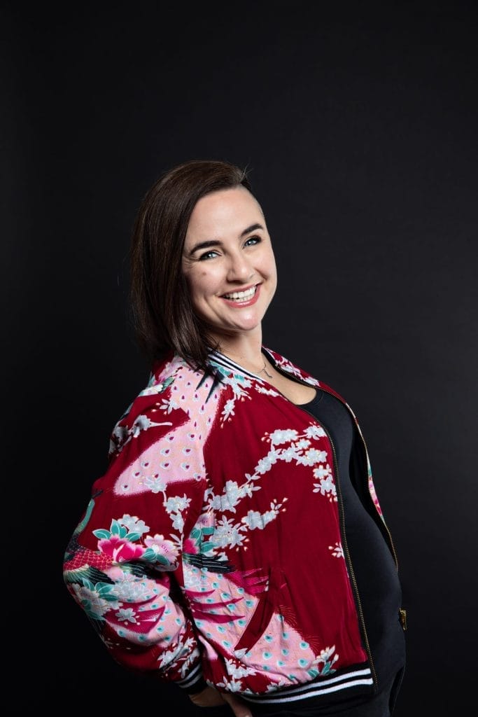 Woman standing against a black background, smiling, wearing a red floral jacket and a black shirt.
