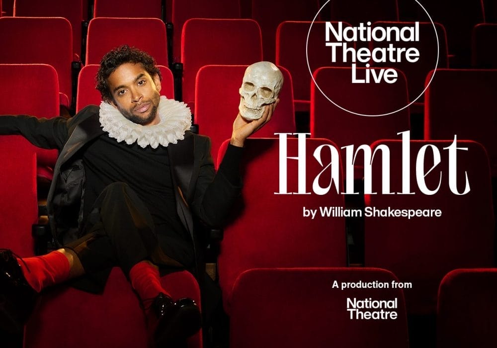 A man with curly dark hair sits in red theatre seats wearing period costume consisting of a black outfit with a white Elizabethan ruff collar. He holds a human skull in his raised right hand, recreating the iconic Hamlet pose. The background shows rows of empty red velvet theatre seats. The National Theatre Live logo appears in a white circle in the upper right corner. Large white text reads "Hamlet" with "by William Shakespeare" below it, and "A production from National Theatre" at the bottom right.