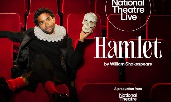 A man with curly dark hair sits in red theatre seats wearing period costume consisting of a black outfit with a white Elizabethan ruff collar. He holds a human skull in his raised right hand, recreating the iconic Hamlet pose. The background shows rows of empty red velvet theatre seats. The National Theatre Live logo appears in a white circle in the upper right corner. Large white text reads "Hamlet" with "by William Shakespeare" below it, and "A production from National Theatre" at the bottom right.