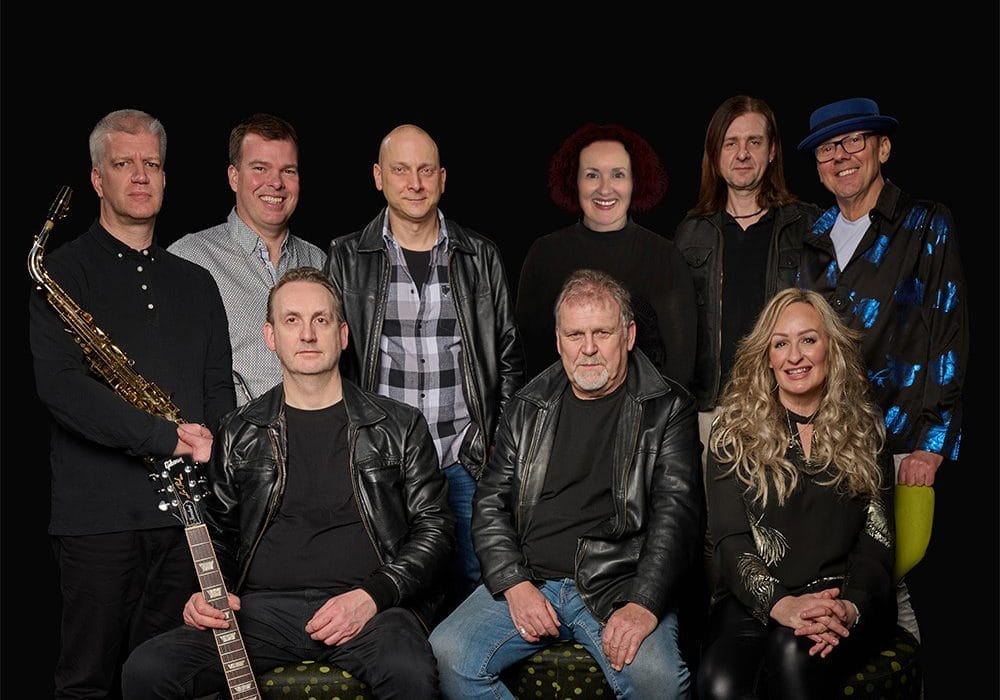 a band, with various instruments visible including a saxophone and guitar. The group consists of about 9-10 people arranged in two rows, with some wearing leather jackets and casual attire.