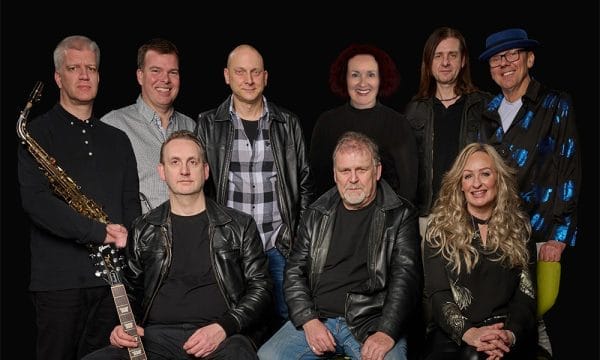 a band, with various instruments visible including a saxophone and guitar. The group consists of about 9-10 people arranged in two rows, with some wearing leather jackets and casual attire.